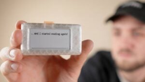 A small 3D-printed e-ink reader resting on a desk beside a USB-C cable and a paperback.